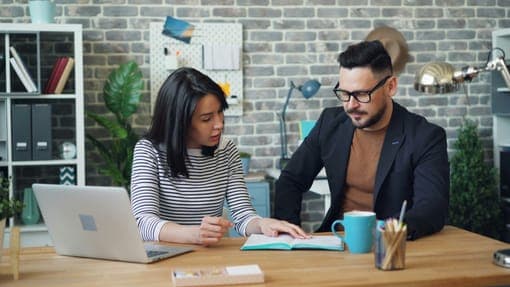 A woman in a striped sweater and a man in a dark blazer talking over a notebook at a desk