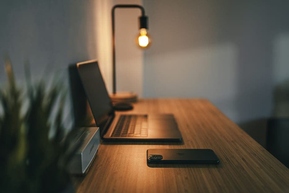 A laptop and a phone on a wooden desk lit by a small lamp