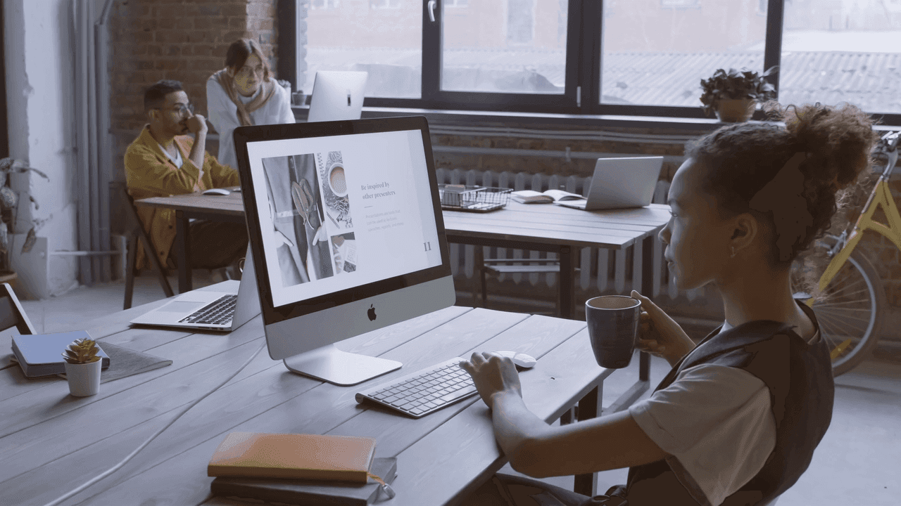 A woman at a long table working at an iMac while colleagues talk behind her