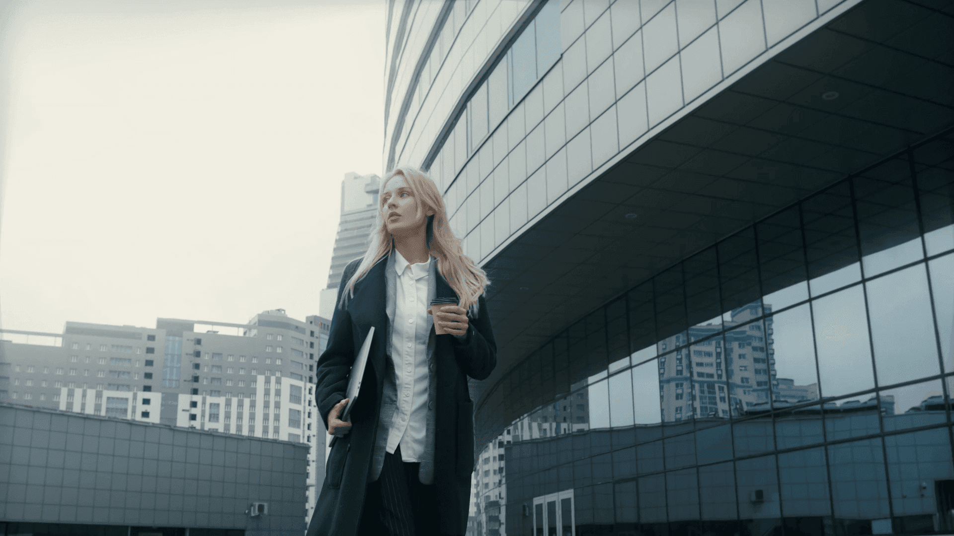 A woman in a dark coat holding a laptop in front of a glass office building