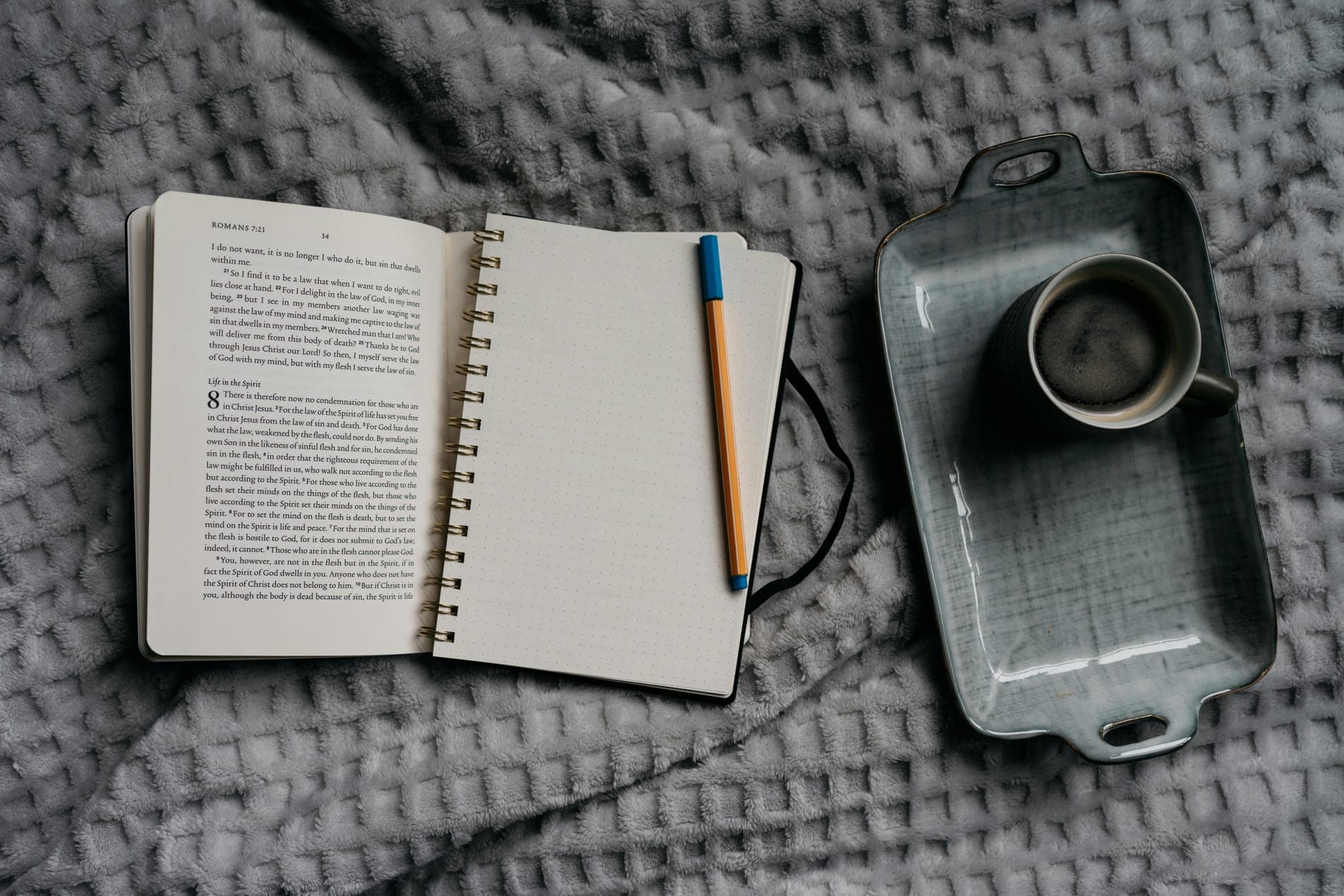 An open notebook, a pen and a cup of coffee on a grey knitted blanket