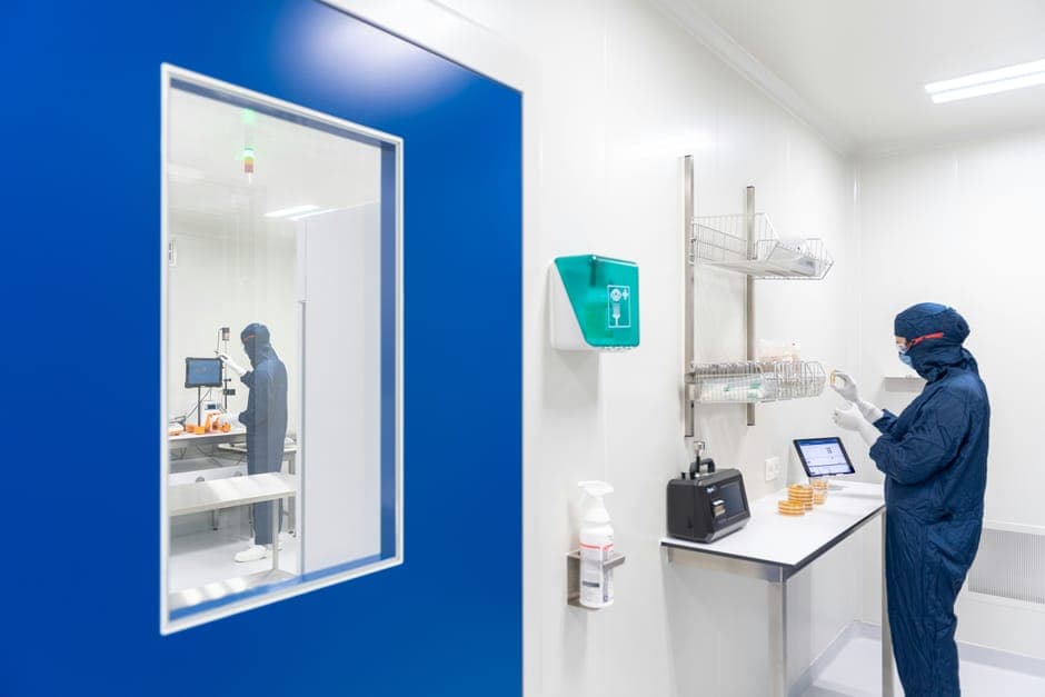 A technician in a blue cleanroom suit working beside a lab window