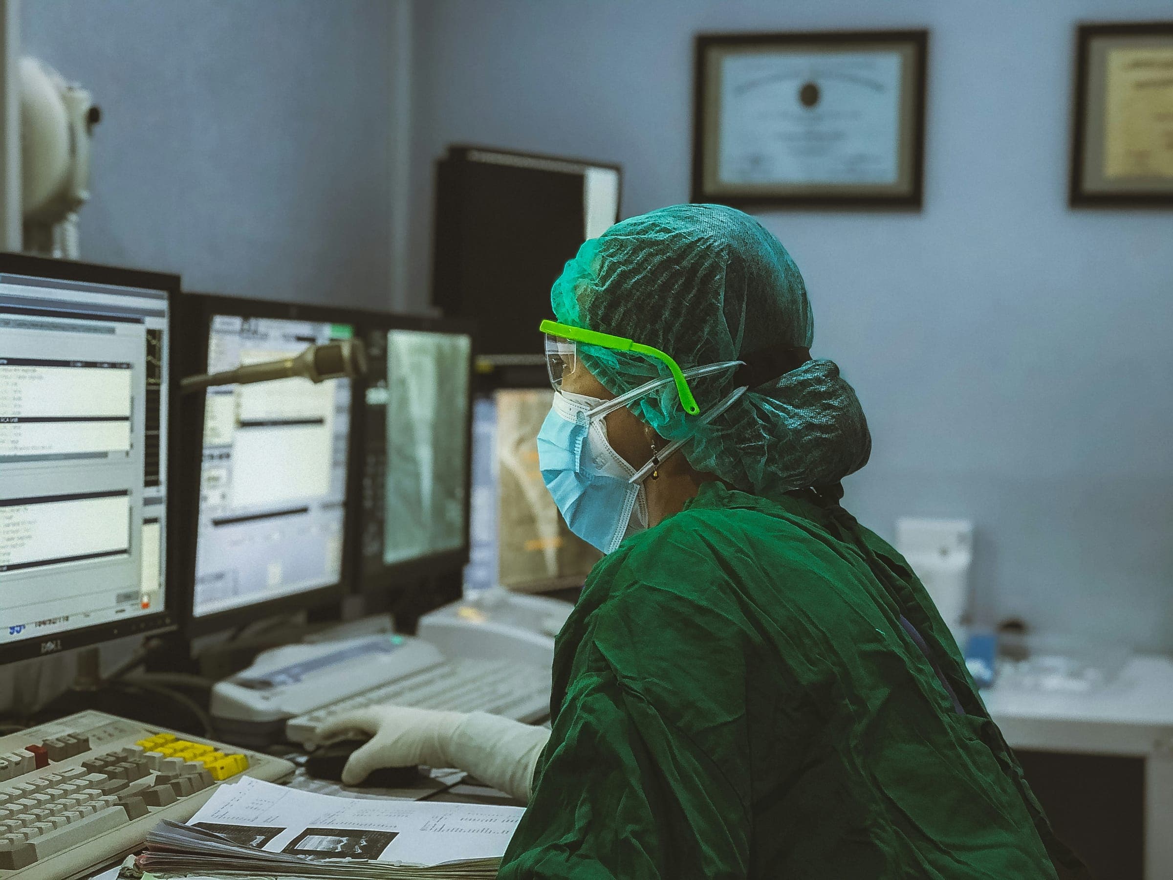 A health worker in green scrubs and a mask checking several monitors