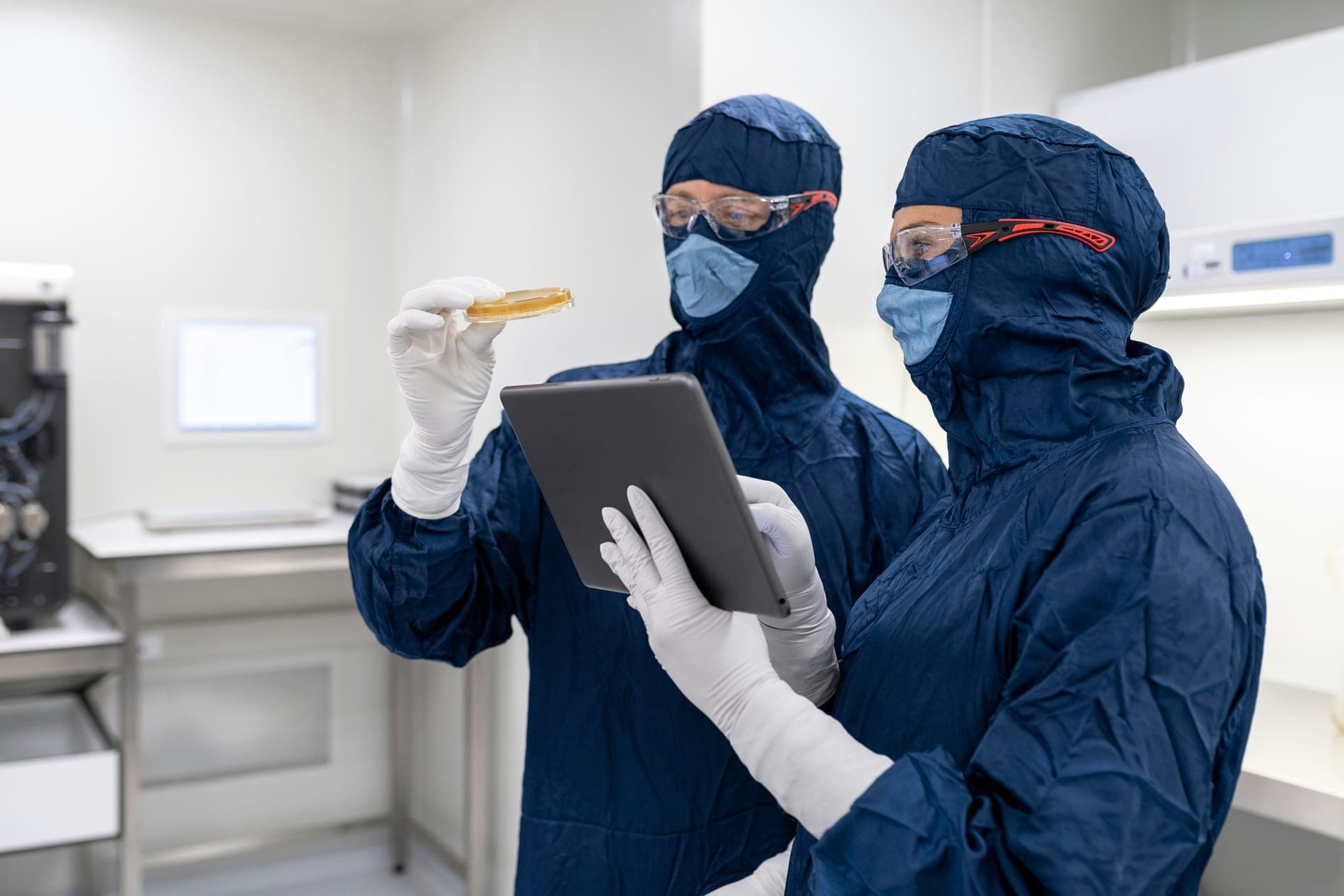 Two lab workers in blue cleanroom suits looking at a tablet
