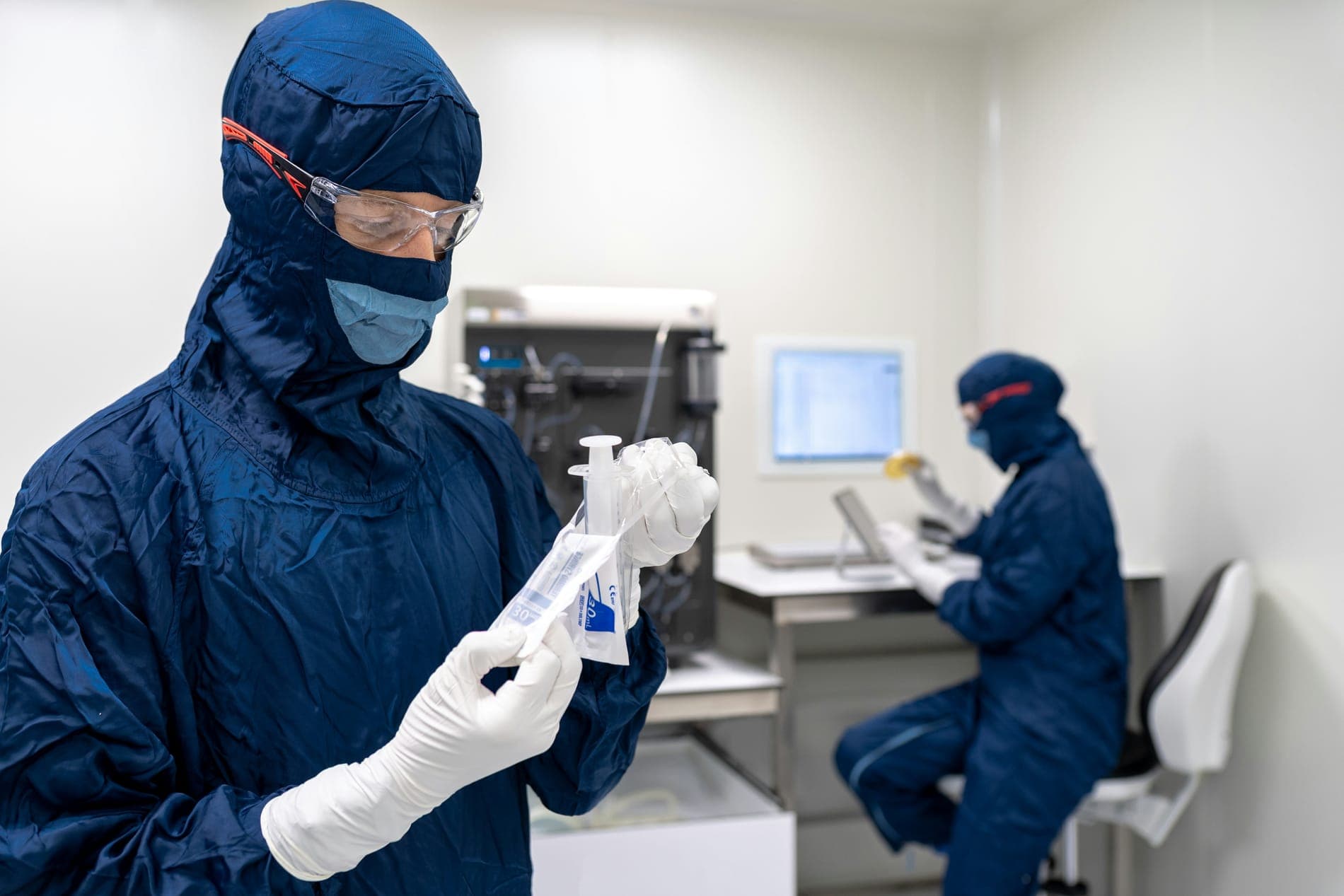 A worker in a blue cleanroom suit holding a sample bottle, a colleague working behind