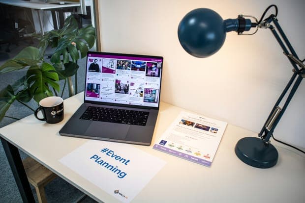 A laptop, a mug and an event planning sign on a desk beside a lamp
