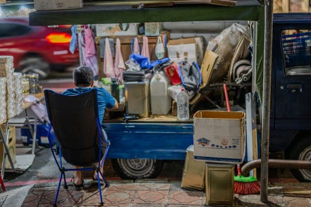 A man sitting in a folding chair beside a blue pickup truck loaded with goods