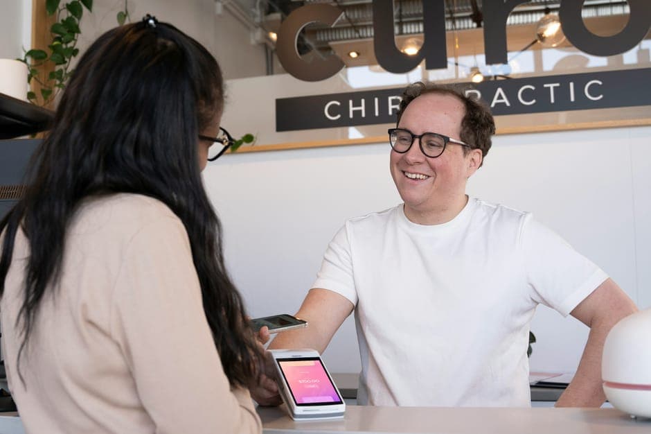 Two people chatting at a chiropractic clinic reception desk with a card reader