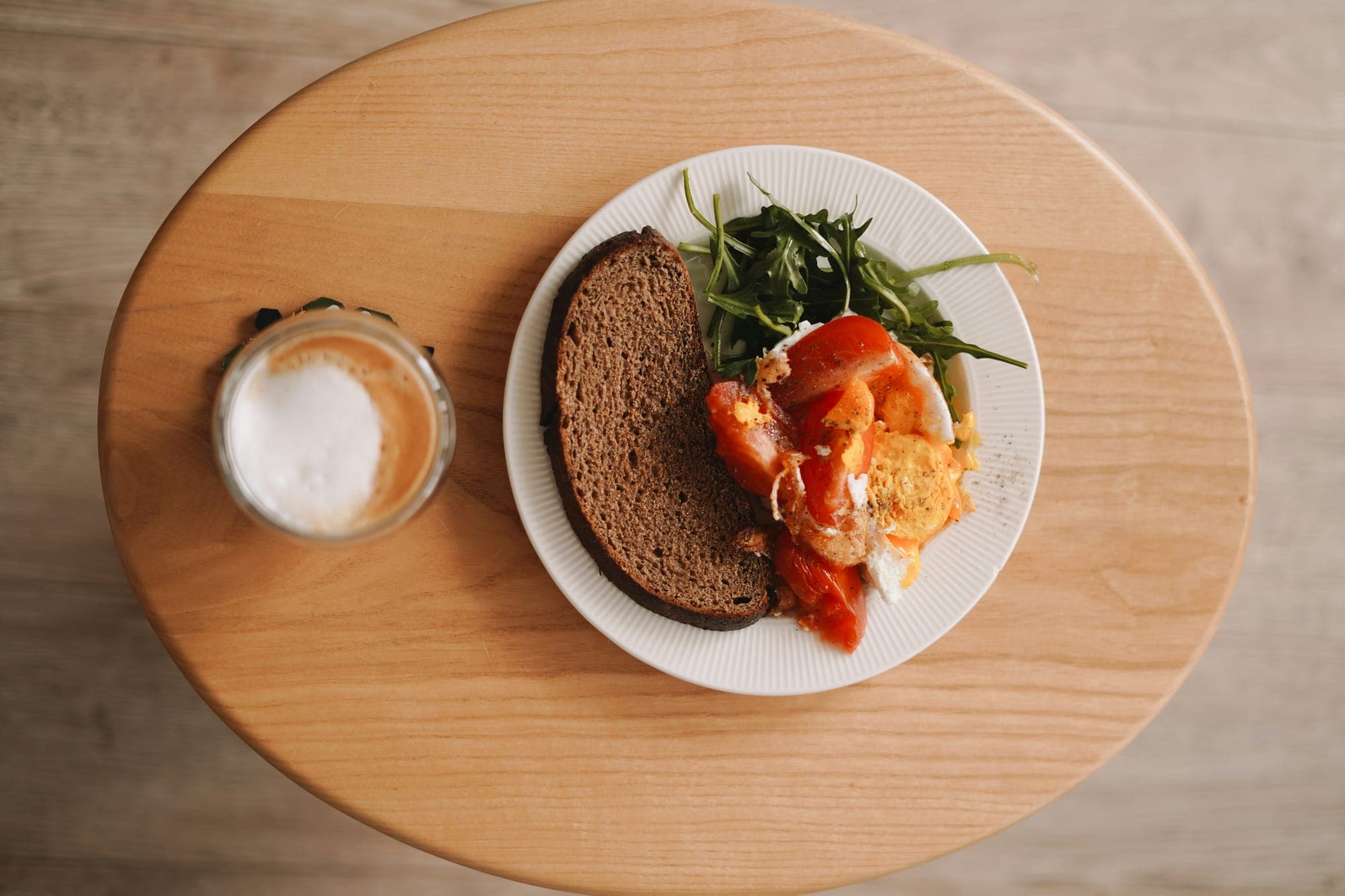 Bread, tomatoes and greens on a plate beside a coffee on a wooden table, seen from above