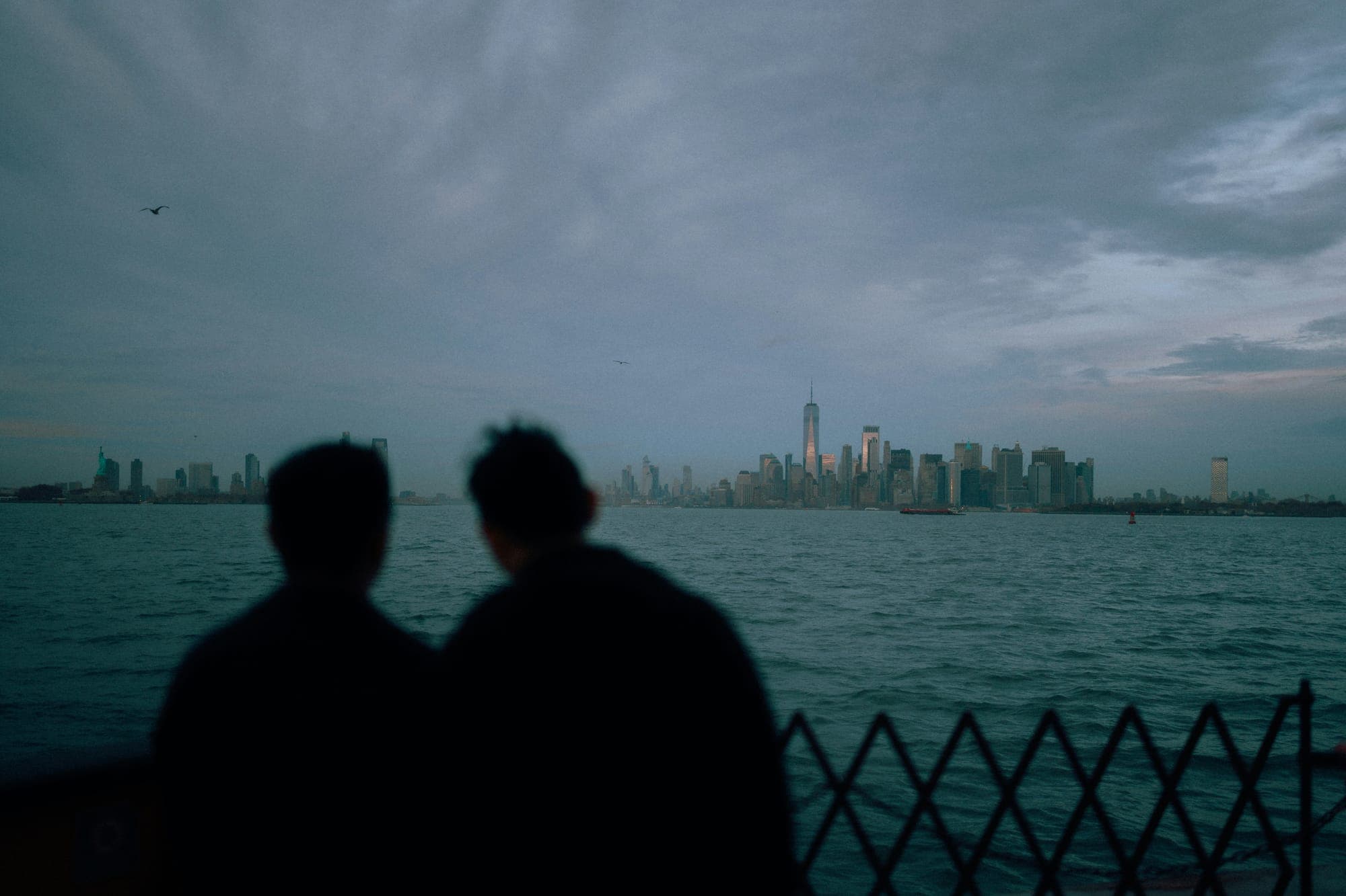 Two silhouettes sitting by the water facing a distant city skyline at dusk