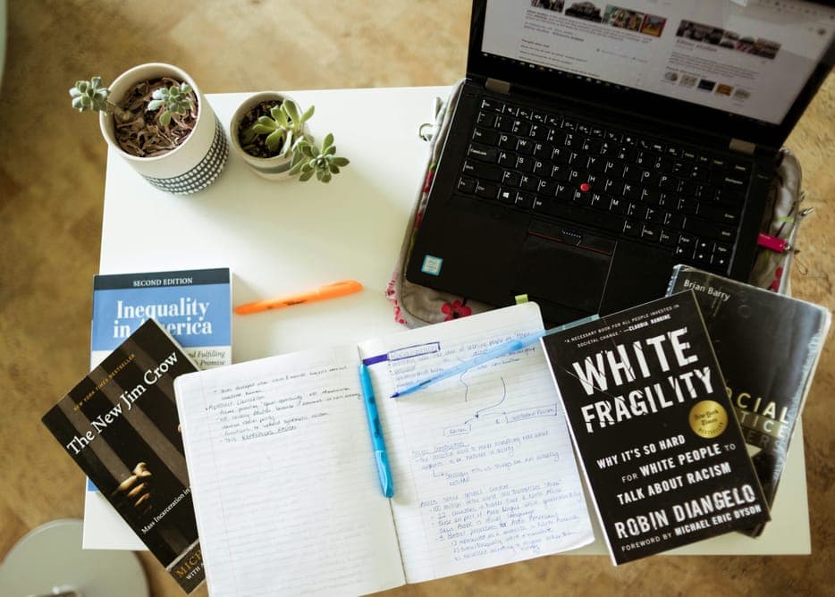 A laptop, an open notebook and books on a white desk seen from above