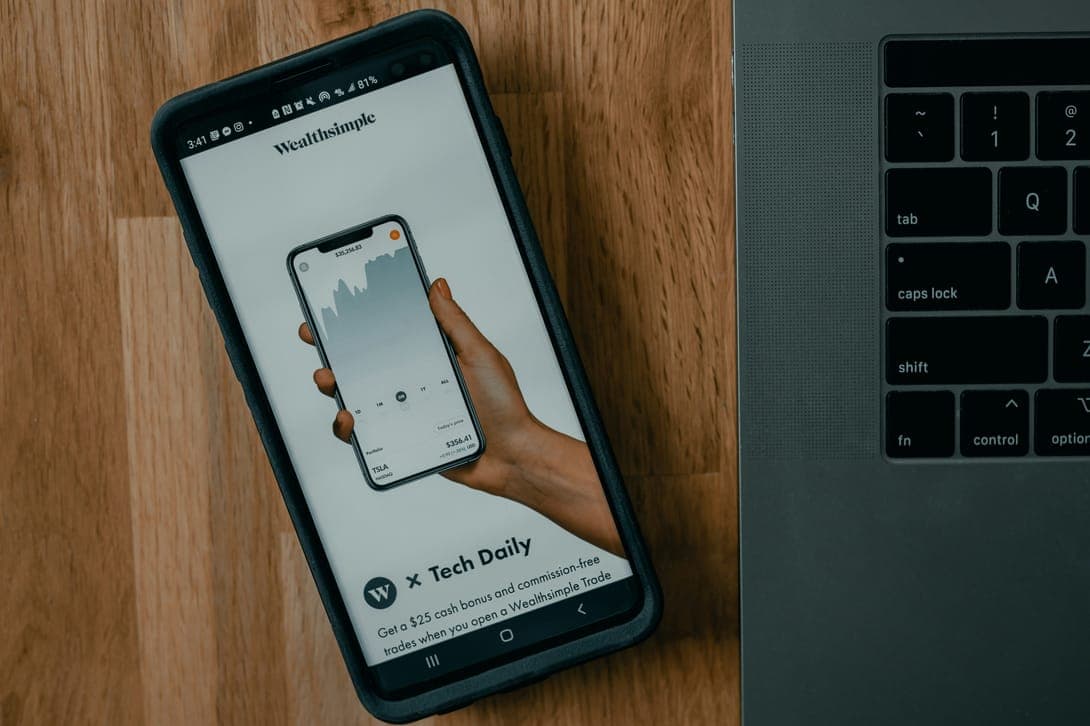 A hand holding a smartphone with a finance app, a laptop keyboard behind it