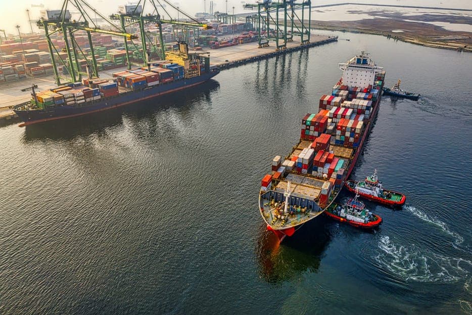 A container ship leaving a port with crane gantries, seen from above