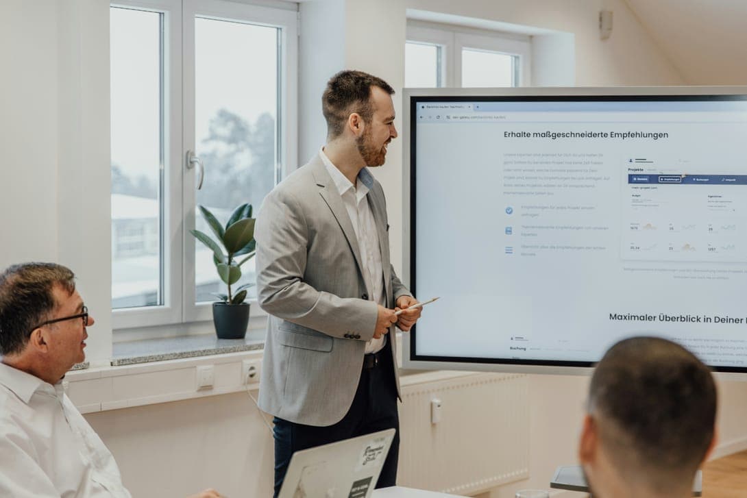 A man in a grey blazer presenting at a wall screen to a seated team
