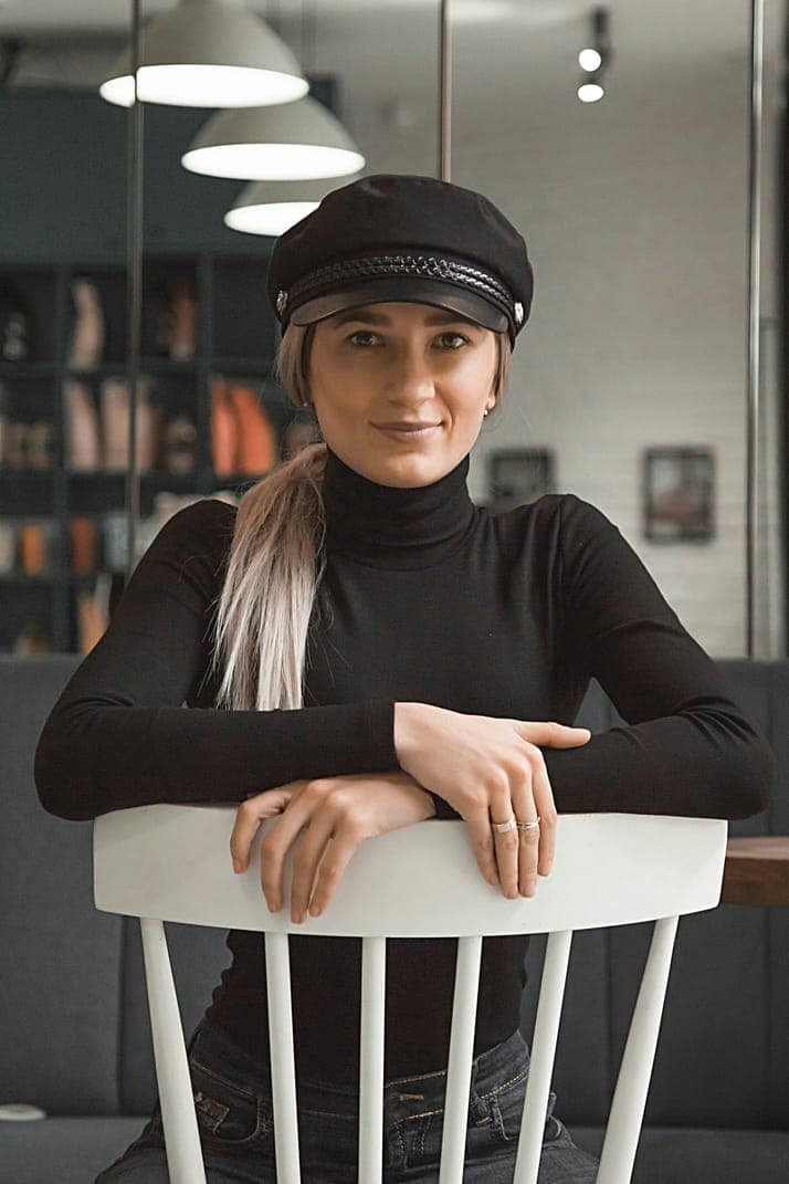 A woman in a black cap and turtleneck leaning on a white chair